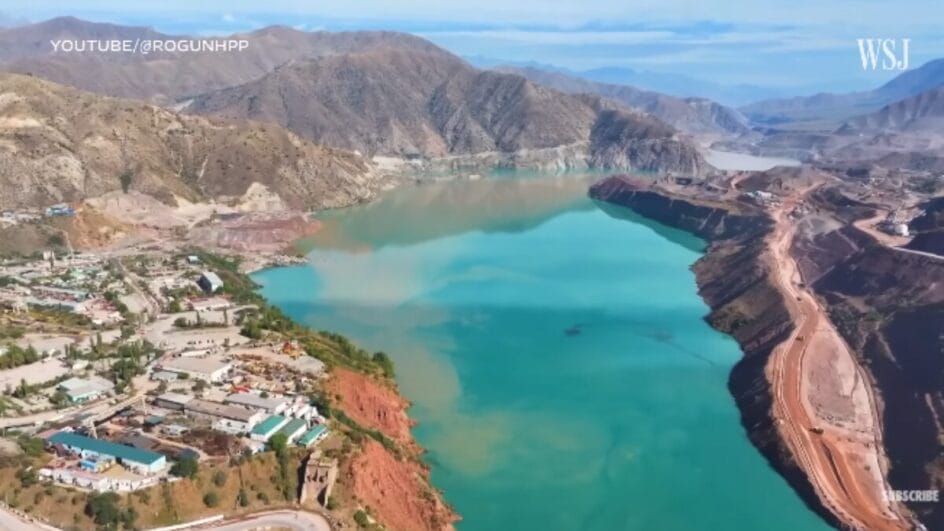 Vista aérea da barragem de Rogun em construção, mostrando um grande reservatório de água turquesa em um vale montanhoso árido.