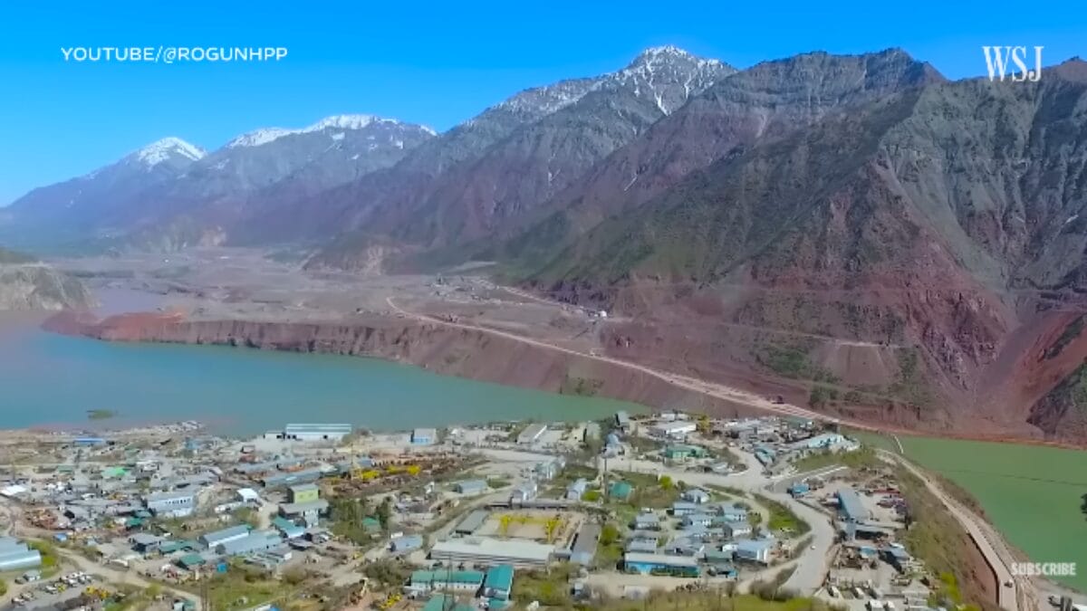 Vista panorâmica da área de construção da barragem de Rogun, com um grande lago verde à esquerda e montanhas rochosas ao fundo.