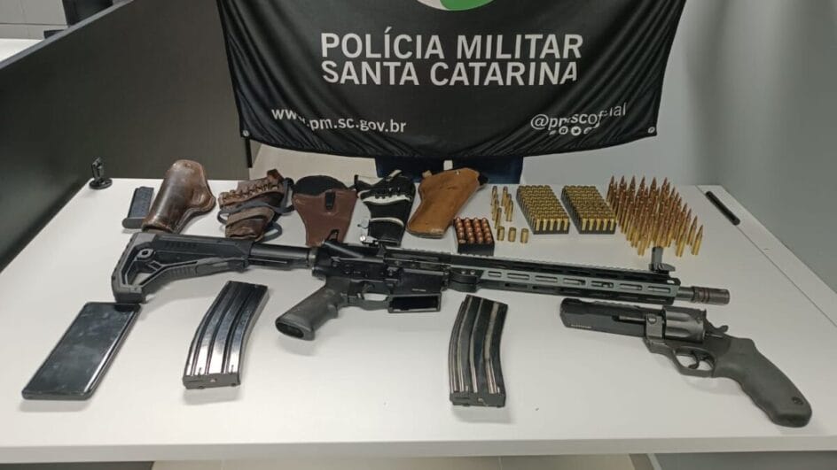 Asenal apreendido em ocorrência de violência contra mulher em Xanxerê. Armas e munições dispostas sobre mesa, com pano ao fundo escrito "Polícia Militar de Santa Catarina"