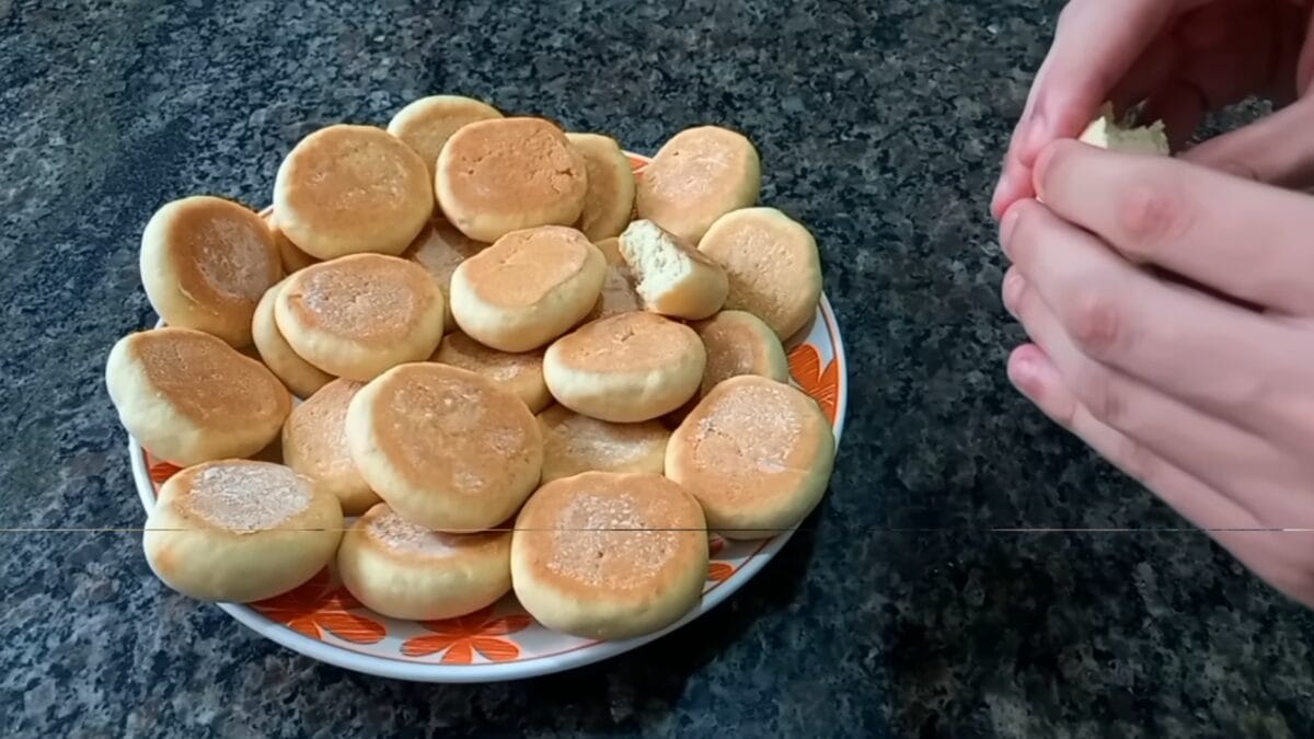 Prato cheio de bolachas caseiras douradas e macias, com uma pessoa mostrando o interior fofo de uma delas.