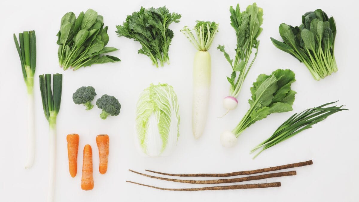 Flat lay (vista superior) de vegetais frescos organizados em fundo branco, comparando espinafre, folhas de nabo, nabo japonês longo (daikon), brócolis, cenouras e cebolinhas.