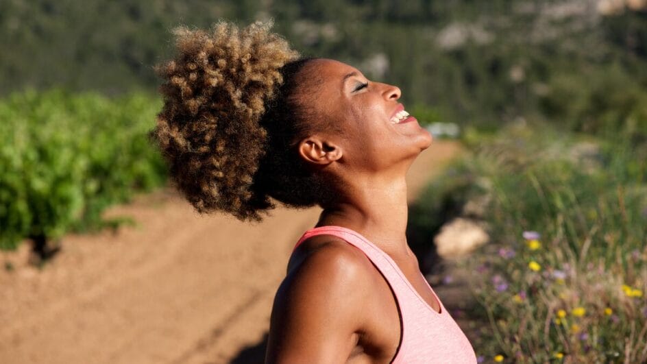 Mulher sorrindo e respirando profundamente durante exercício ao ar livre, simbolizando saúde mental e alívio do estresse.