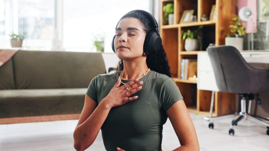 Mulher meditando com fones de ouvido em casa, prática de respiração e mindfulness para reduzir ansiedade.