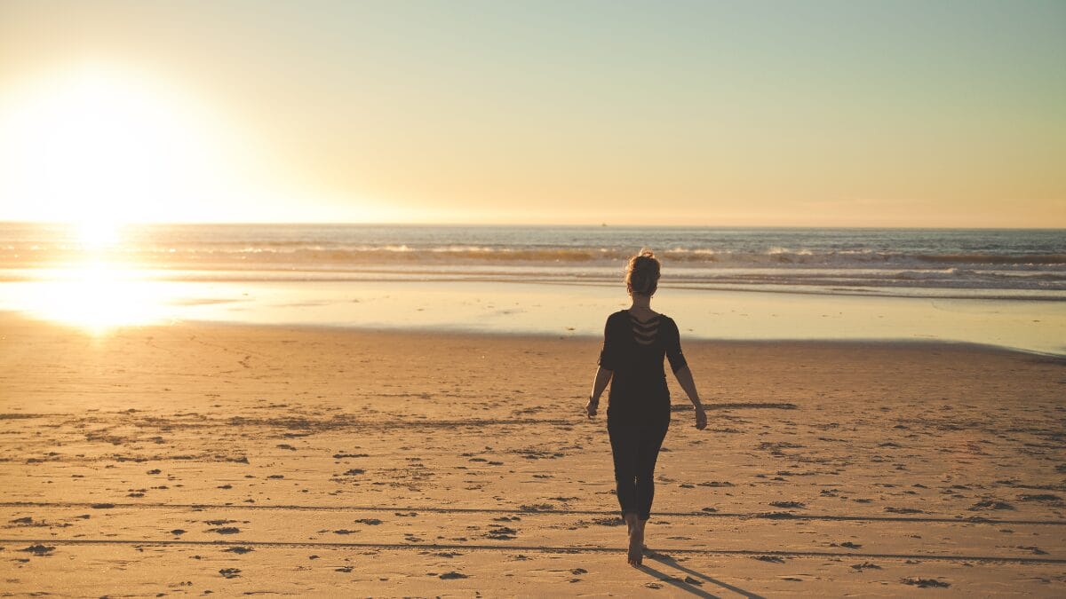 Pessoa caminhando sozinha pela areia da praia ao pôr do sol, com o mar ao fundo.