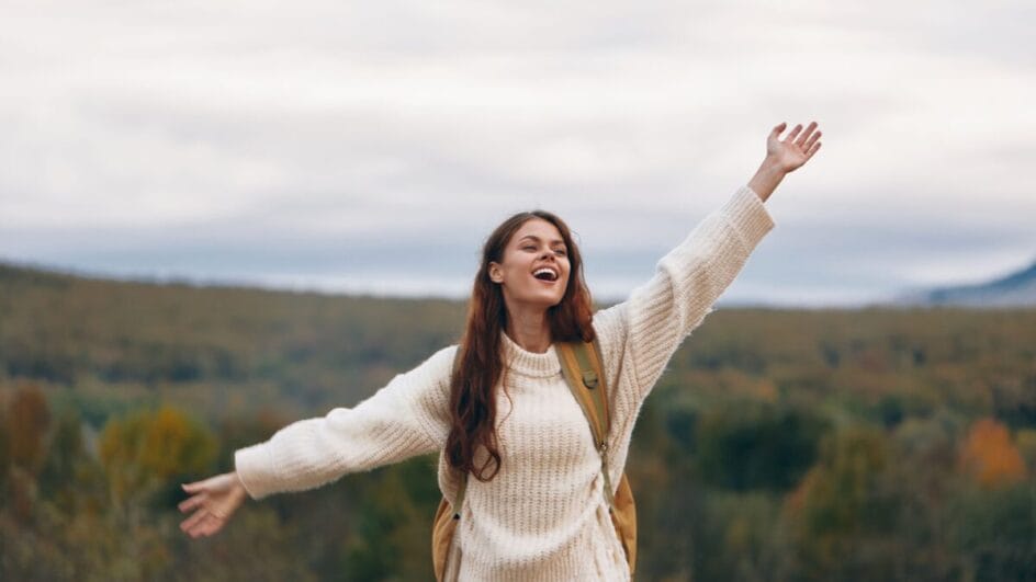Mulher sorrindo com os braços abertos em trilha, sensação de liberdade e bem-estar emocional.