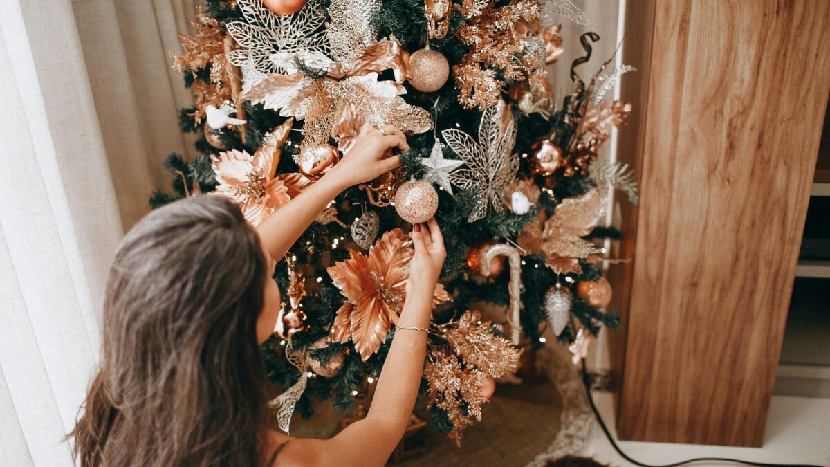 Pessoa decorando uma árvore de Natal com enfeites dourados em ambiente interno.