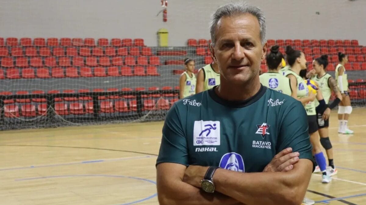 Marco Antônio Queiroga posando para foto com os braços cruzados e camisa do Mackenzie e o time treinando no fundo