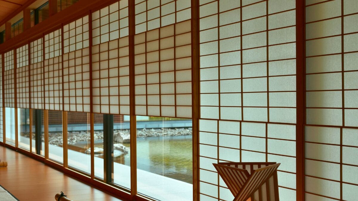 Amplo corredor com piso de madeira polida e uma longa extens&atilde;o de pain&eacute;is shoji. Alguns pain&eacute;is est&atilde;o abertos, permitindo a vis&atilde;o de um espelho d'&aacute;gua e pedras na &aacute;rea externa, integrando a arquitetura com a &aacute;gua.
