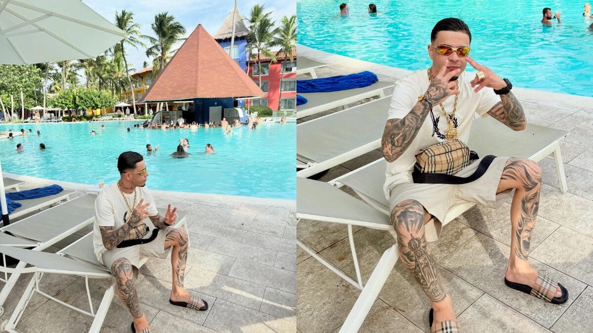 MC Tuto sentado em uma espregui&ccedil;adeira em um resort de luxo. Ele veste camiseta branca, bermuda clara, chinelos e bolsa da Burberry. Ao fundo, uma grande piscina com bar molhado em formato de pir&acirc;mide.