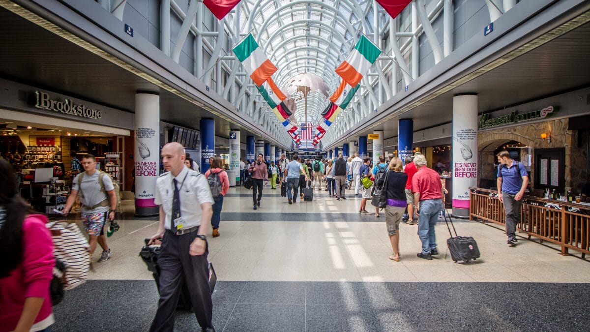 Foto ampla de um corredor movimentado no Aeroporto O'Hare. O teto é alto, com claraboias arqueadas, e decorado com dezenas de bandeiras de diferentes países penduradas em duas fileiras. Ao fundo, uma estrutura esférica prateada pende do teto. Passageiros caminham com malas pelo corredor central, ladeados por lojas e restaurantes, incluindo uma placa da "Brookstone".