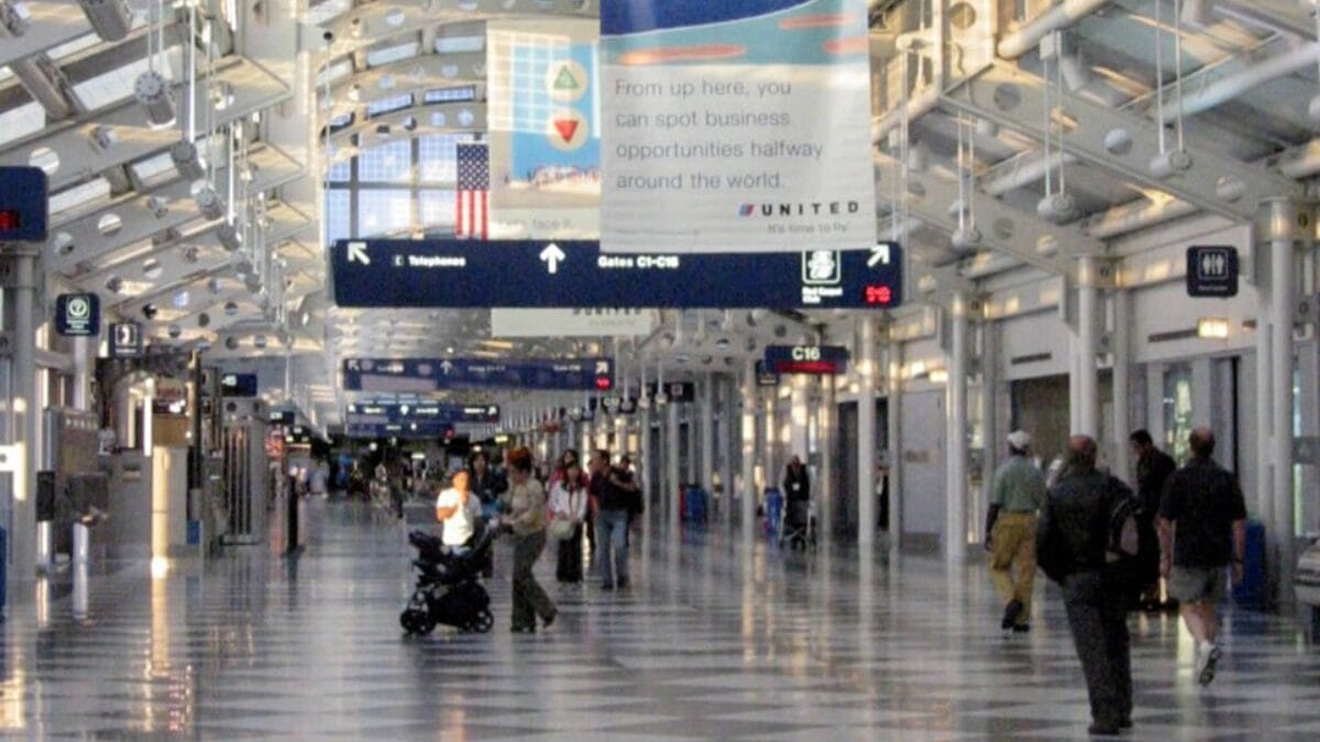Corredor de embarque moderno e iluminado no Terminal 1. A estrutura do teto é composta por vigas de aço branco com design industrial e janelas superiores que deixam entrar luz natural. Placas de sinalização azul indicam "Portões C1-C18" e publicidade da United Airlines está suspensa. Pessoas caminham calmamente pelo piso polido que reflete as luzes.
