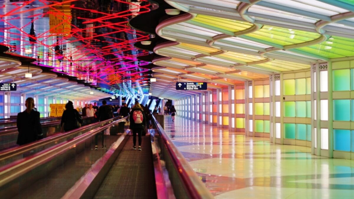 Túnel subterrâneo futurista que conecta saguões. O teto possui uma escultura de luz neon ondulada e multicolorida que reflete intensamente no teto espelhado e no chão. As paredes laterais são painéis de vidro em tons pastéis iluminados. Viajantes utilizam as esteiras rolantes em ambos os lados, cercados pelas luzes vibrantes.