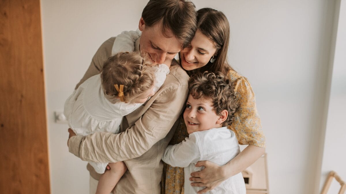 Grupo familiar se abraça em um ambiente interno acolhedor. Dois adultos envolvem duas crianças em um abraço coletivo, transmitindo carinho, proteção e vínculo emocional.