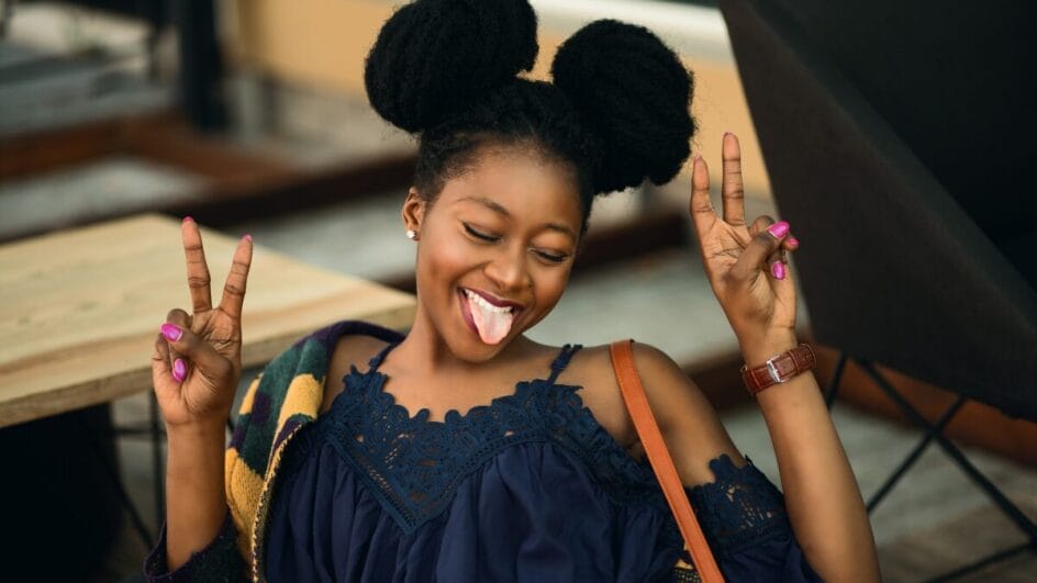 Pessoa sentada em um ambiente urbano descontraído faz o gesto de sinal de paz com as duas mãos. A roupa casual e a postura relaxada transmitem alegria, leveza e espontaneidade.