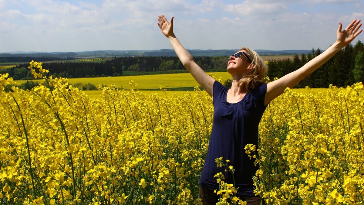 Pessoa em um vasto campo de flores amarelas abre os braços para o céu. A paisagem ampla e ensolarada transmite sensação de liberdade, renovação e alegria ao ar livre.