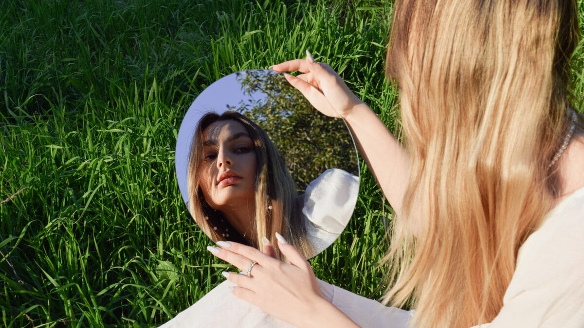 Pessoa segurando um espelho circular em meio à grama verde, refletindo parcialmente o céu e a vegetação ao redor. A composição sugere auto-observação, identidade e diálogo entre o corpo e o ambiente natural.