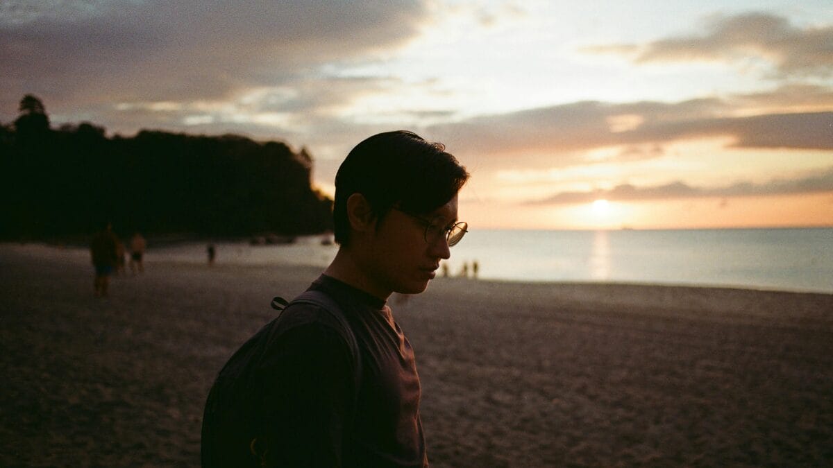 Pessoa caminhando sozinha pela praia ao entardecer, com o sol baixo iluminando a areia e o mar ao fundo. A cena passa sensação de tranquilidade, reflexão e encerramento de um ciclo do dia.