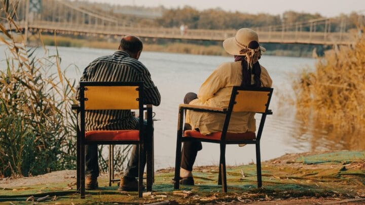 Duas pessoas sentam-se em cadeiras à beira de um rio, observando a água e uma ponte ao fundo. A cena sugere conversa tranquila, descanso e compartilhamento de tempo em um ambiente natural.