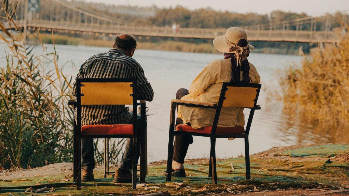 Duas pessoas sentam-se em cadeiras à beira de um rio, observando a água e uma ponte ao fundo. A cena sugere conversa tranquila, descanso e compartilhamento de tempo em um ambiente natural.