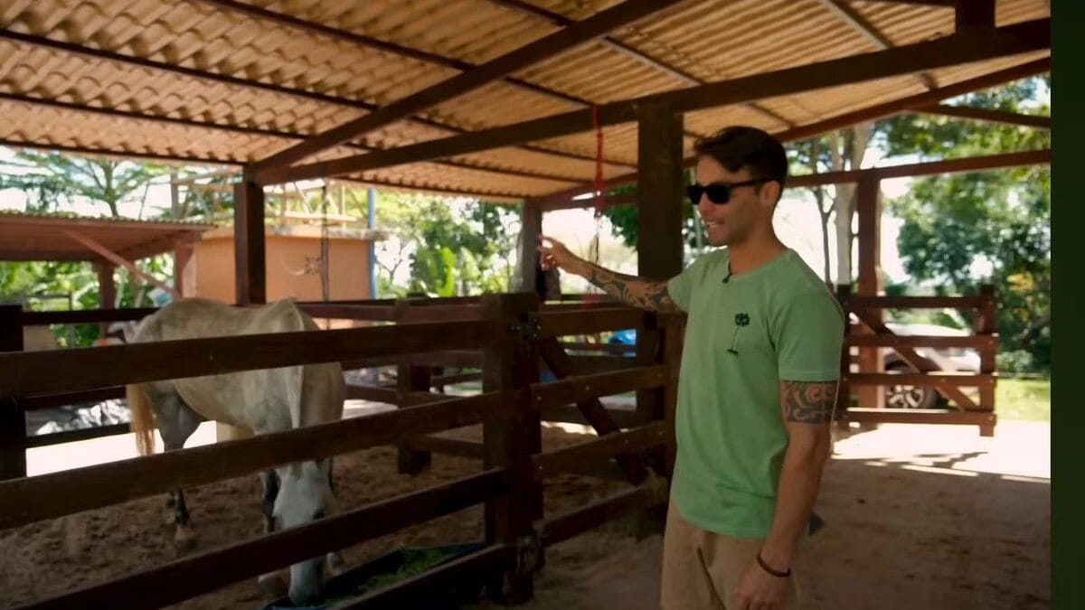 Dentro de um est&aacute;bulo de madeira com teto coberto, Daniel Cady aponta para um cavalo branco que est&aacute; comendo em um cocho de alimenta&ccedil;&atilde;o.