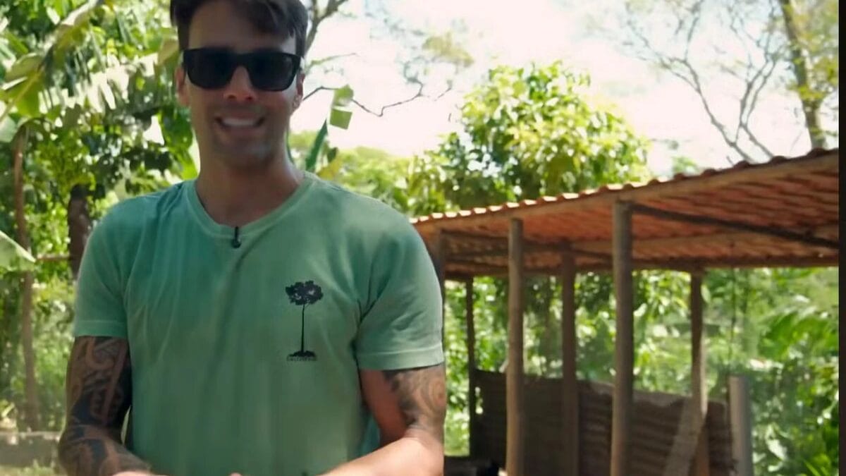 Daniel Cady, usando &oacute;culos escuros e camiseta verde-clara que mostra uma tatuagem tribal no bra&ccedil;o direito, sorri e gesticula em meio a uma vegeta&ccedil;&atilde;o tropical densa e exuberante em seu s&iacute;tio sob um c&eacute;u azul ensolarado.