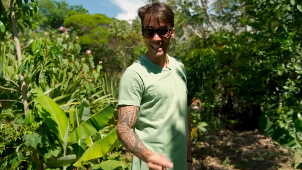 Daniel Cady, nutricionista, usa &oacute;culos escuros e uma camisa verde-&aacute;gua, gesticulando e sorrindo em meio a uma vegeta&ccedil;&atilde;o tropical densa e exuberante em seu s&iacute;tio.