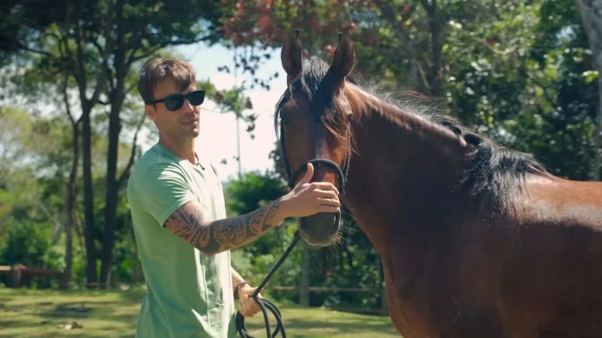 Daniel Cady, usando &oacute;culos escuros e camisa verde, acaricia suavemente o focinho de um cavalo marrom em um ambiente rural ensolarado com &aacute;rvores ao fundo.