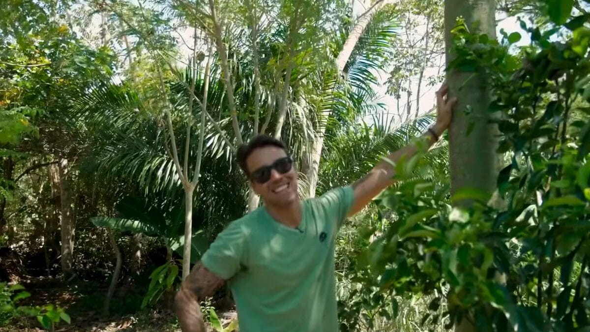 Daniel Cady sorri para a c&acirc;mera enquanto se apoia no tronco de uma &aacute;rvore, cercado por folhagem verde e densa em uma &aacute;rea de mata do seu s&iacute;tio.