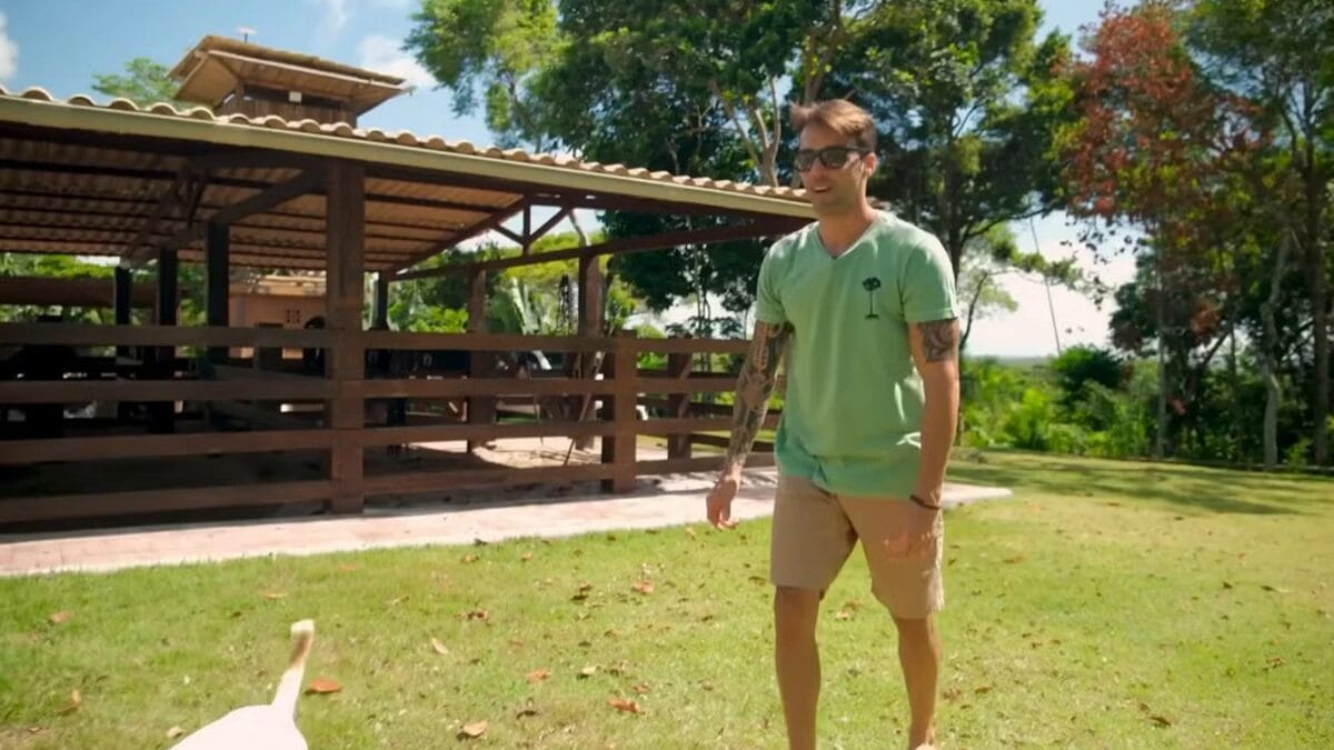 Daniel Cady, de corpo inteiro, est&aacute; em p&eacute; em um gramado verde, olhando para algo fora do quadro. Ao fundo, h&aacute; um grande galp&atilde;o de madeira r&uacute;stica com telhado de telhas.