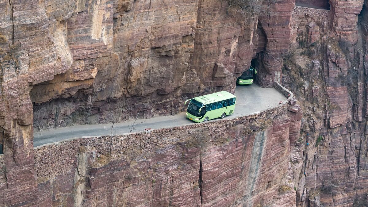 Vista externa da encosta da montanha onde se v&ecirc; dois &ocirc;nibus de turismo verdes percorrendo o caminho estreito esculpido dentro do pared&atilde;o rochoso.