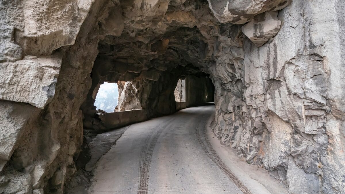 Vista interna de um t&uacute;nel r&uacute;stico e escuro, com paredes de rocha bruta e irregular. H&aacute; aberturas laterais que funcionam como janelas naturais para o precip&iacute;cio.