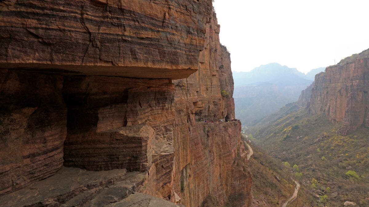 Fotografia de um enorme pared&atilde;o de rocha avermelhada e estratificada. No meio da encosta vertical, nota-se uma linha estreita de estrada esculpida onde pessoas caminham.