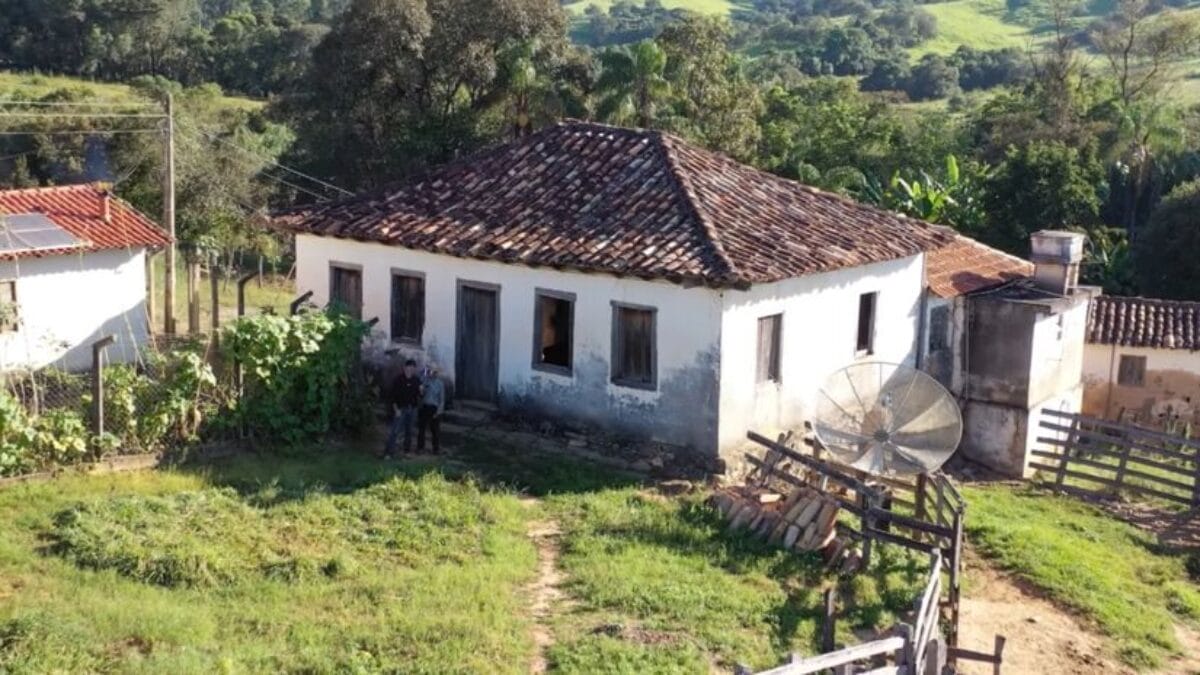 Fotografia a&eacute;rea de uma casa antiga de paredes brancas e telhado de barro envelhecido. Dois dos irm&atilde;os aparecem em p&eacute; na frente da casa, pr&oacute;ximos a uma antena parab&oacute;lica e cercas de madeira. O fundo mostra colinas verdes e &aacute;rvores.