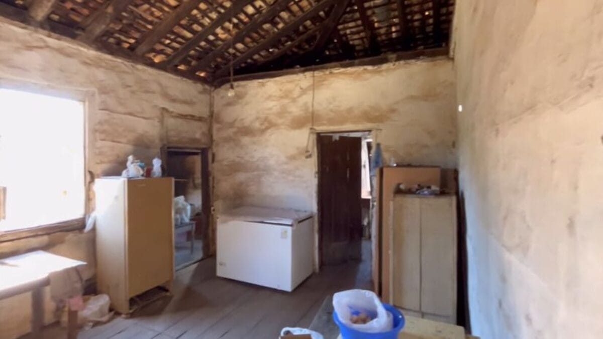 Vista interna de um c&ocirc;modo r&uacute;stico com paredes de barro descascadas e teto de madeira aparente. H&aacute; um freezer branco no centro, arm&aacute;rios de madeira simples e uma janela iluminada &agrave; esquerda.