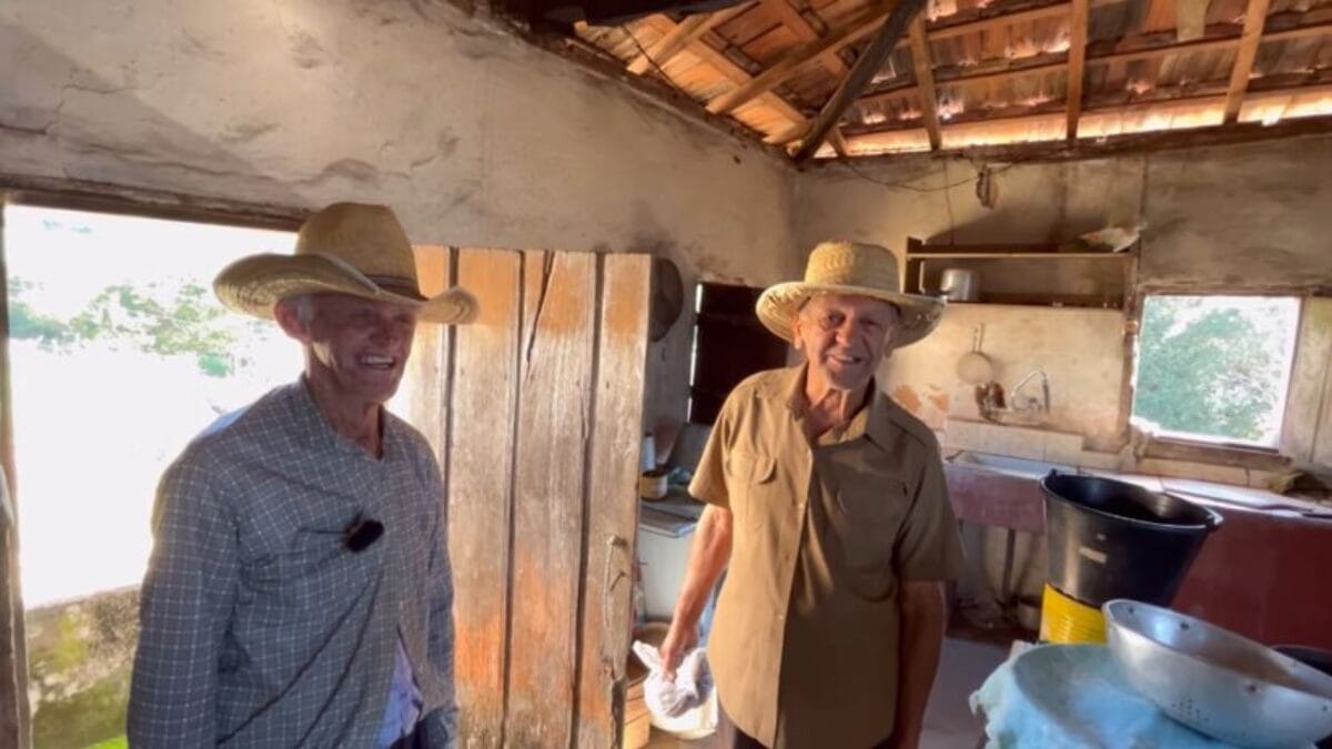 Dois senhores sorridentes usando chap&eacute;us de palha e camisas de bot&atilde;o dentro de uma cozinha r&uacute;stica. O ambiente ao fundo mostra utens&iacute;lios simples e uma janela que d&aacute; para a vegeta&ccedil;&atilde;o externa.