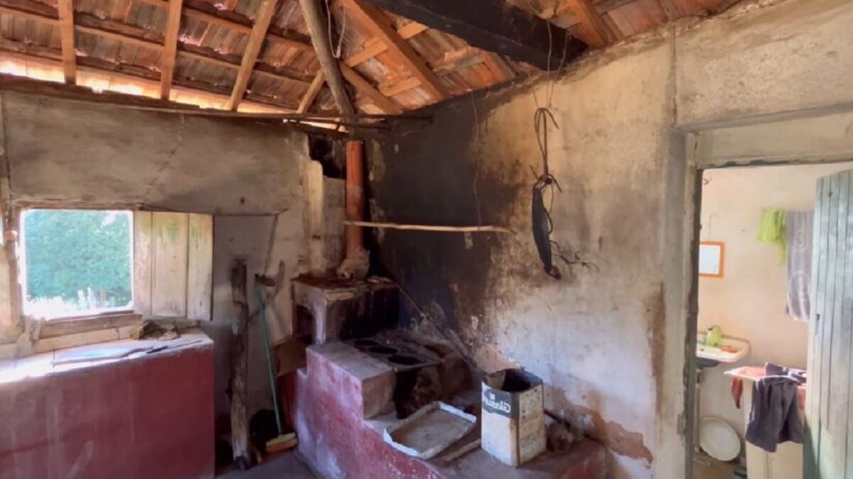 Detalhe de uma cozinha antiga com um fog&atilde;o a lenha de alvenaria pintado de vermelho. As paredes est&atilde;o escurecidas pela fuligem do fogo e o telhado de telhas de barro est&aacute; aparente. Uma porta aberta ao fundo revela um banheiro simples.