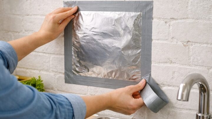 Uma pessoa aplica um quadrado de papel alumínio em uma parede de tijolos brancos acima de uma pia de cozinha, prendendo as bordas com fita adesiva cinza (silver tape).