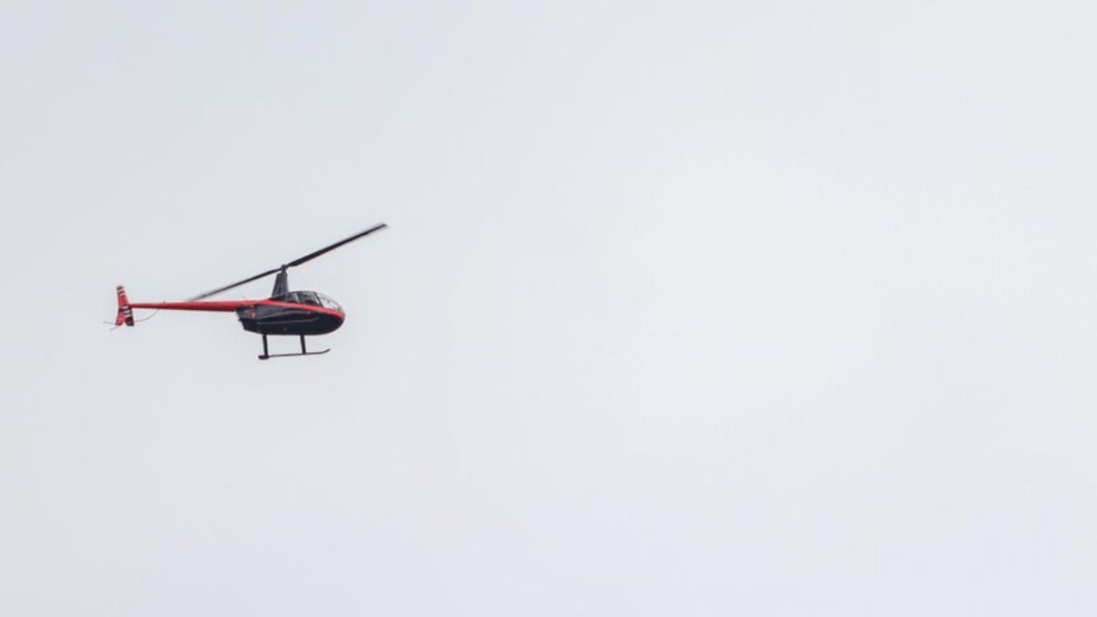 Um helic&oacute;ptero de pequeno porte voando contra um c&eacute;u claro e p&aacute;lido, representando a opera&ccedil;&atilde;o de transporte de troncos.