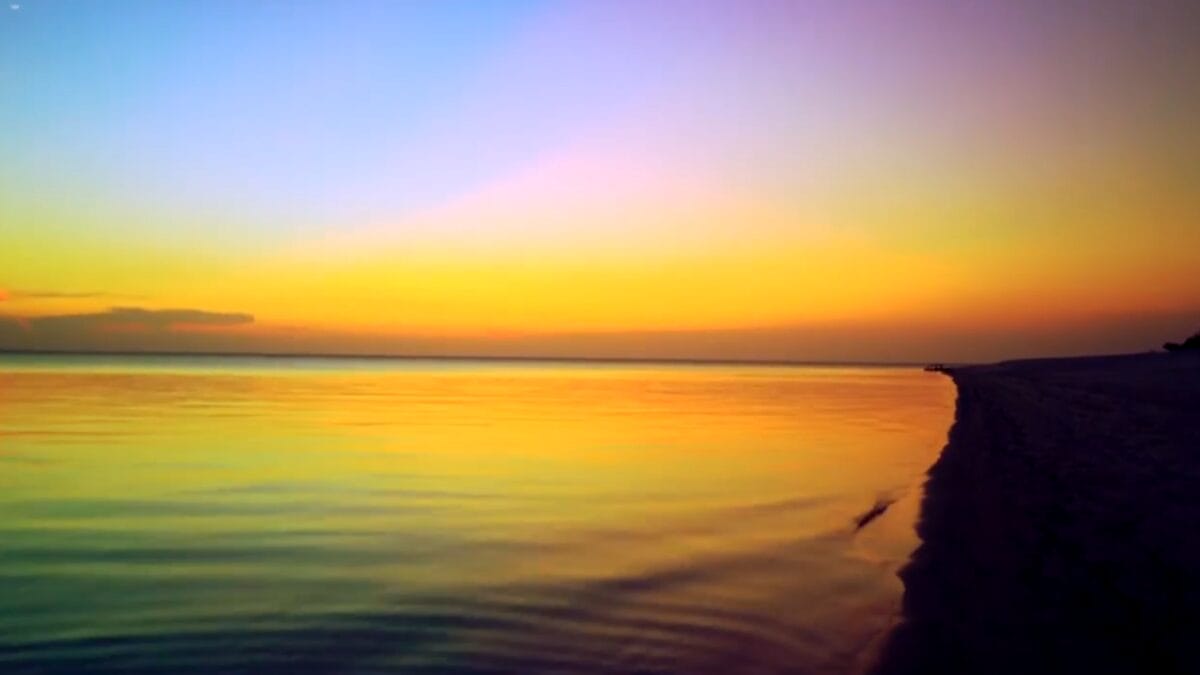 Fotografia ao nível da areia de uma praia fluvial na Amazônia durante o pôr do sol, com águas calmas refletindo tons de laranja, amarelo e azul.