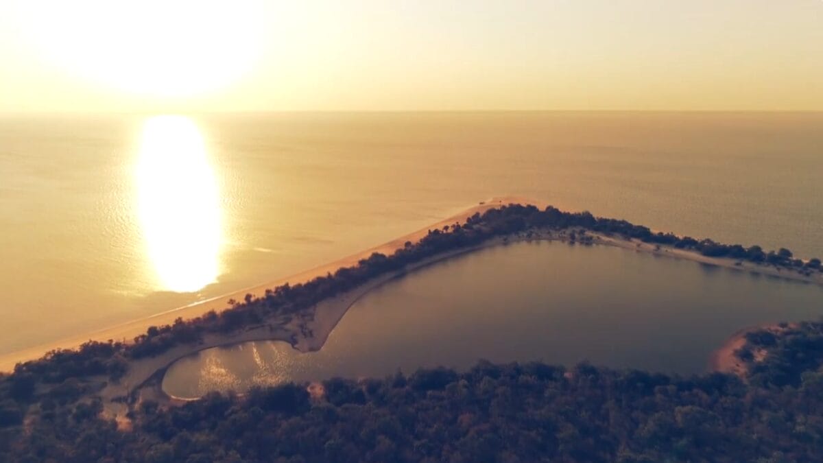 Vista aérea de uma península de areia formando uma lagoa interna na Amazônia, com o sol refletindo intensamente na água ao fundo.