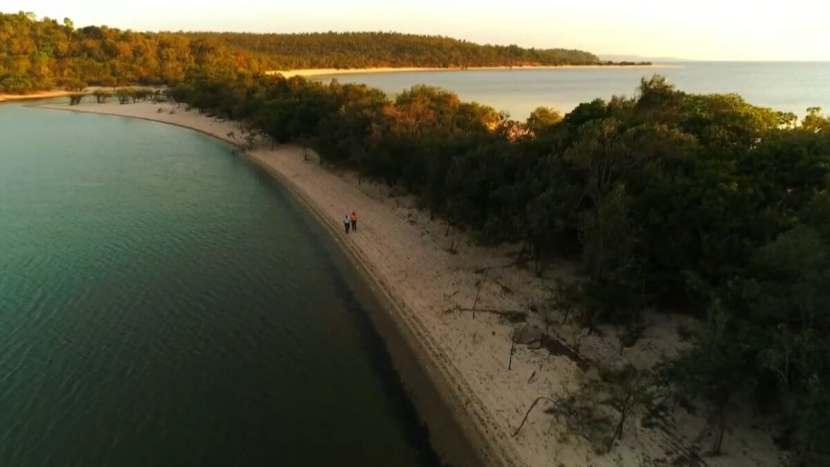 Vista aérea diagonal de duas pessoas caminhando por uma praia de areia clara entre a densa Floresta Amazônica e as águas do rio.
