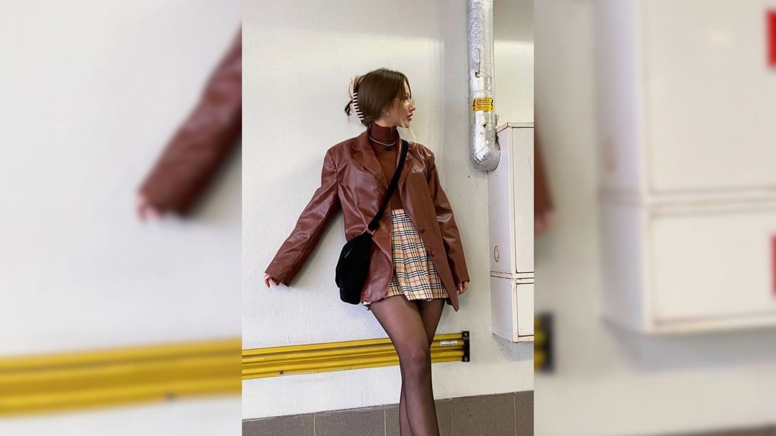 Mulher encostada na parede usando blazer marrom de couro com saia xadrez curta e meia-calça preta, em look de outono urbano.