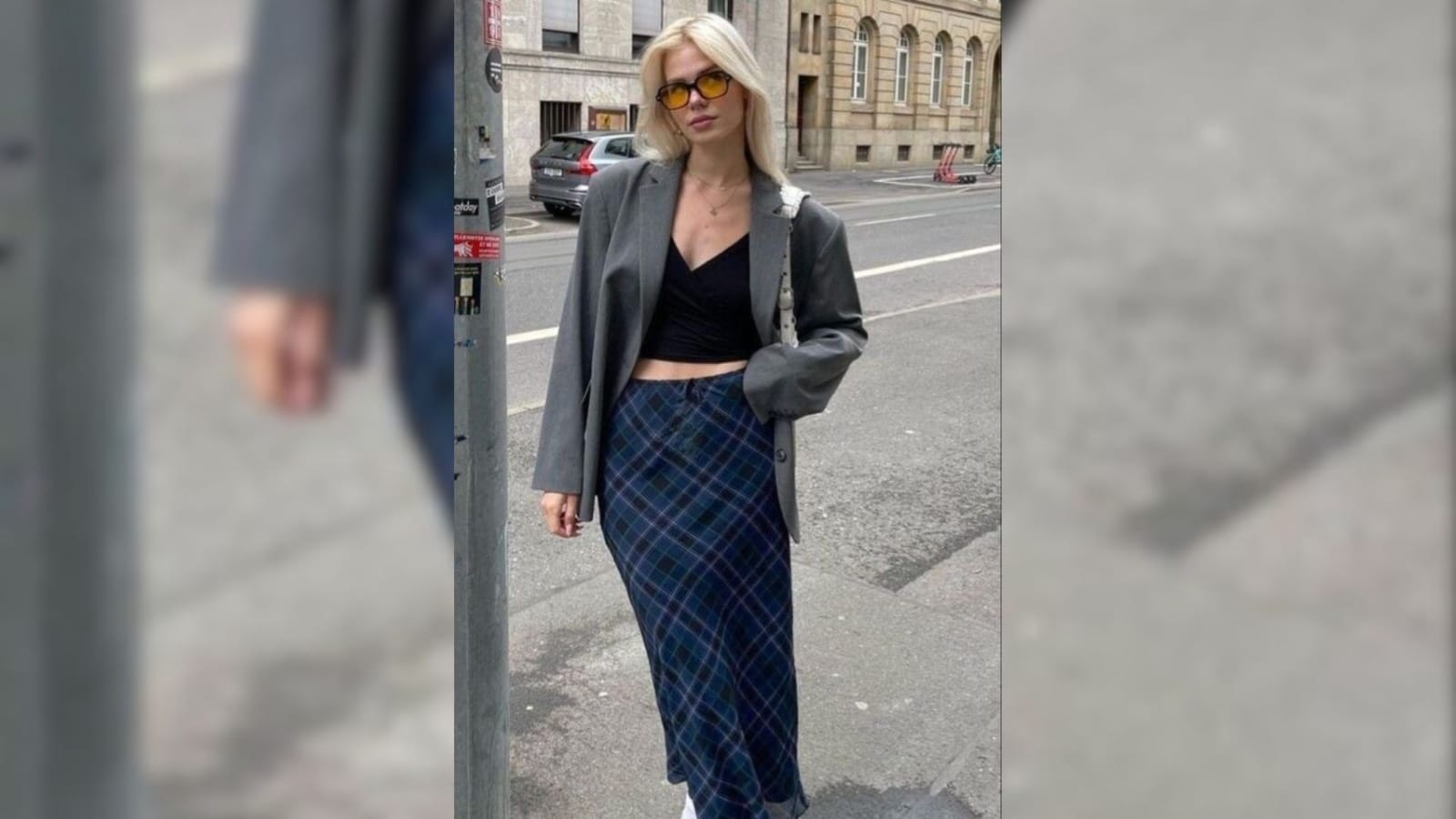Mulher loira usando blazer cinza oversized, top preto e saia midi xadrez azul em ambiente urbano.