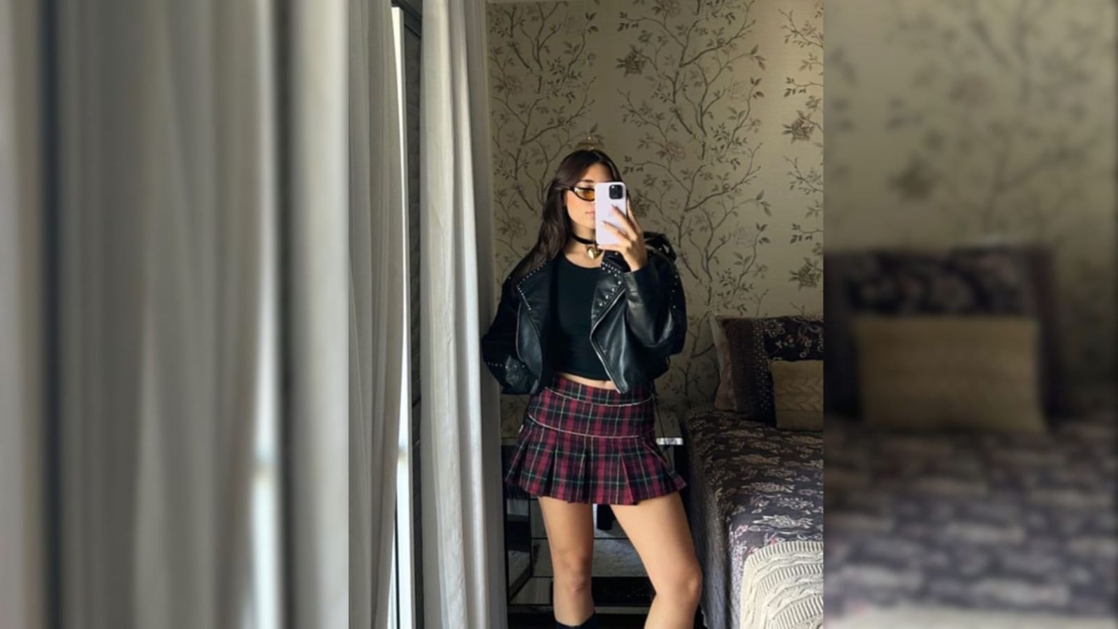 Mulher tirando selfie com jaqueta de couro preta e saia xadrez curta em tons escuros, em ambiente interno.