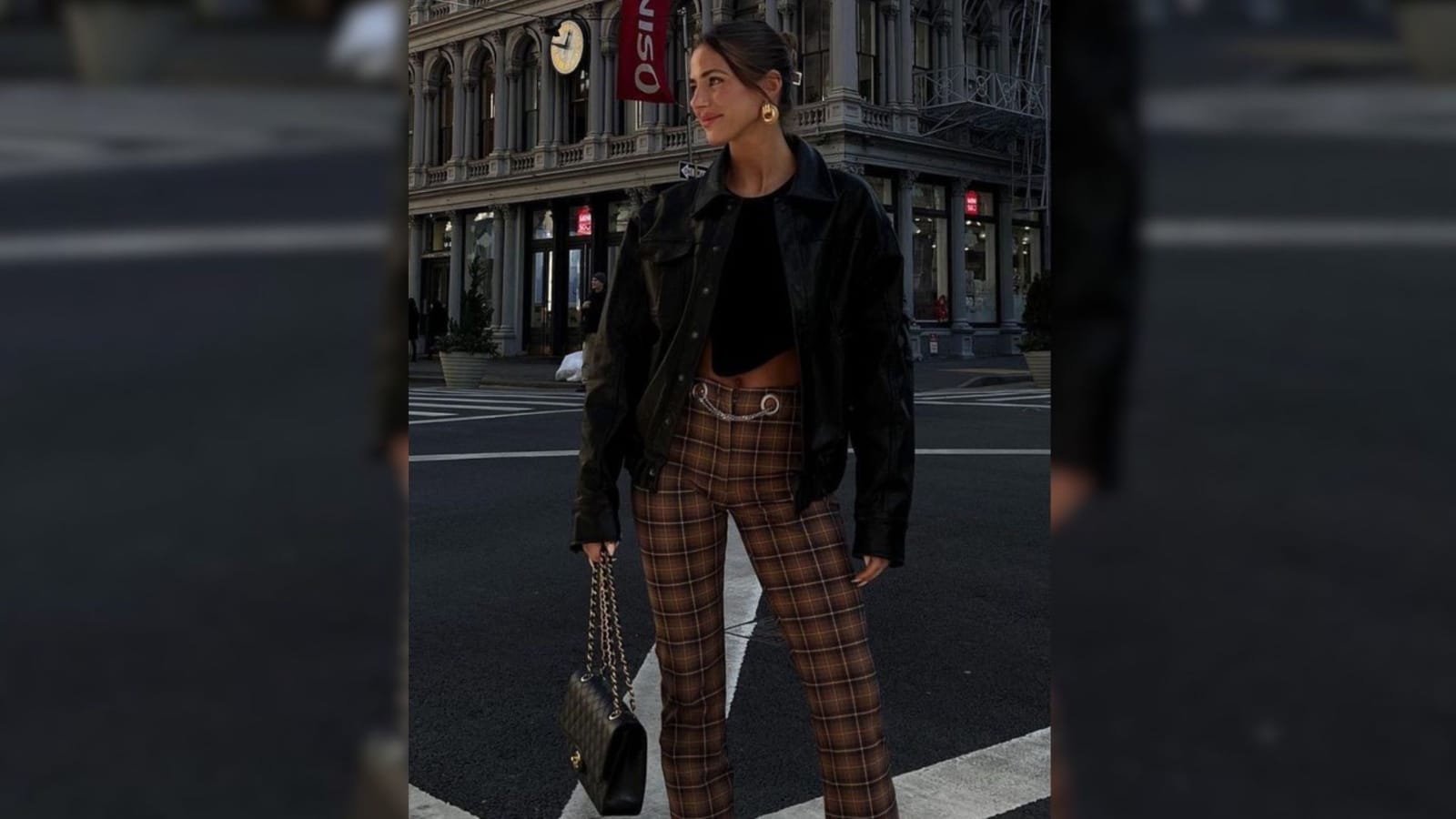 Mulher em rua urbana usando calça xadrez marrom, jaqueta preta e bolsa de corrente.