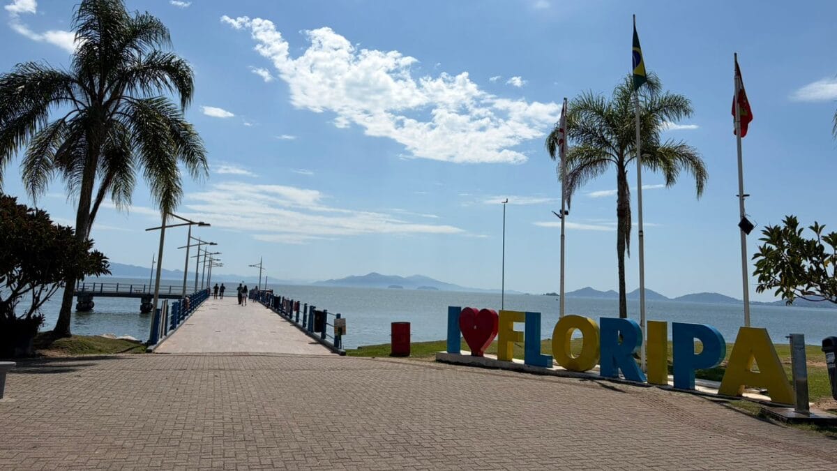 Trapiche da Beira-Mar Norte e letreiro &ldquo;Eu amo Floripa&rdquo;, com pedestres caminhando e a ba&iacute;a de Florian&oacute;polis ao fundo.