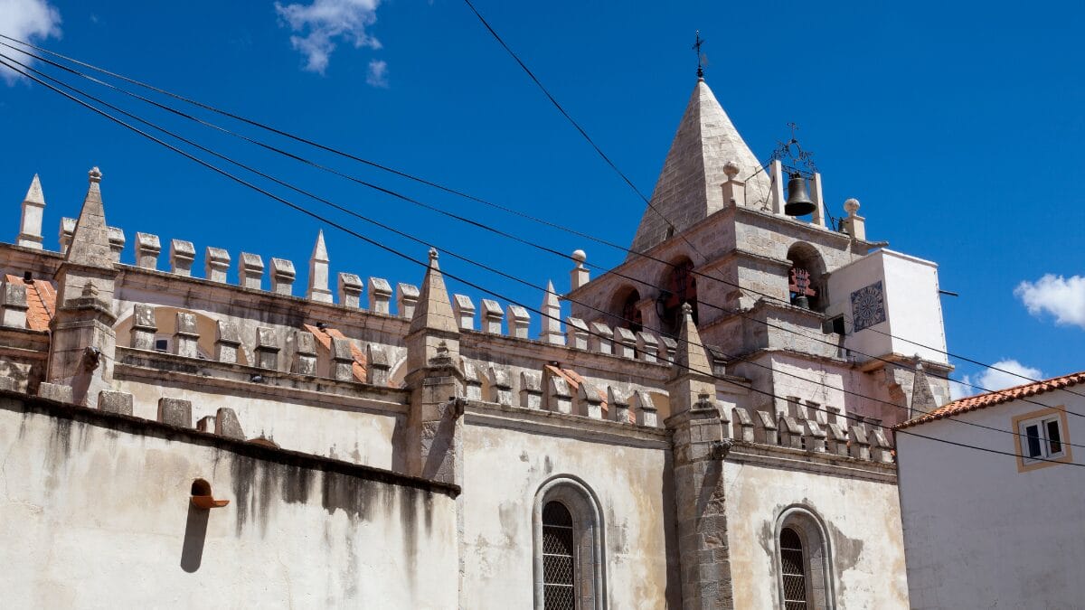 Fachada lateral da Igreja de Nossa Senhora da Assun&ccedil;&atilde;o em Elvas, apresentando detalhes manuelinos, ameias no topo e uma torre sineira pontiaguda contra um c&eacute;u azul.