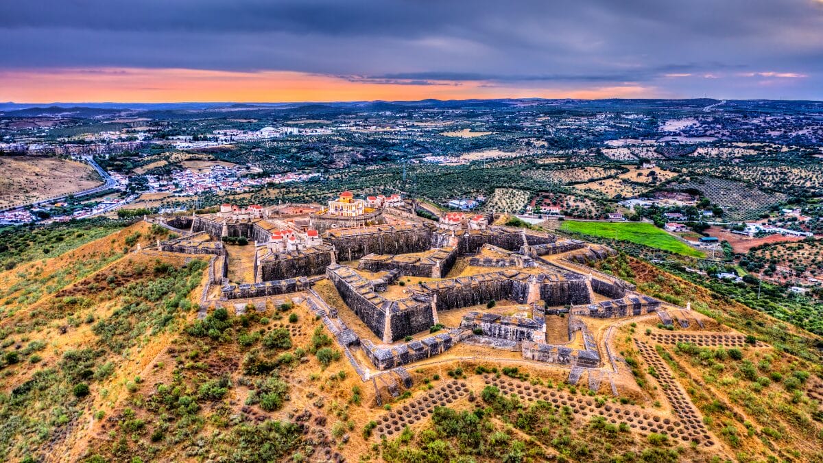 Fotografia a&eacute;rea do Forte de Nossa Senhora da Gra&ccedil;a em Elvas sob um c&eacute;u de fim de tarde. A estrutura em formato de estrela destaca-se no topo de uma colina cercada por vegeta&ccedil;&atilde;o.