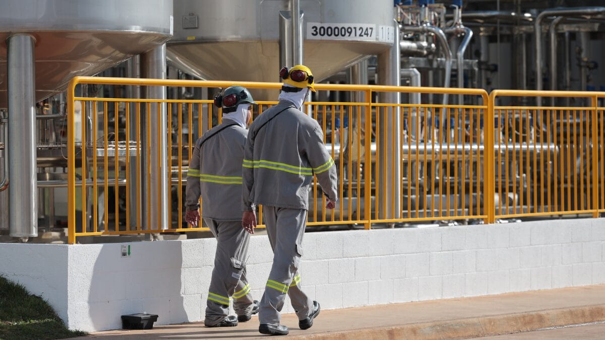 Dois trabalhadores caminham por &aacute;rea industrial com tanques met&aacute;licos e tubula&ccedil;&otilde;es em f&aacute;brica de latic&iacute;nios.