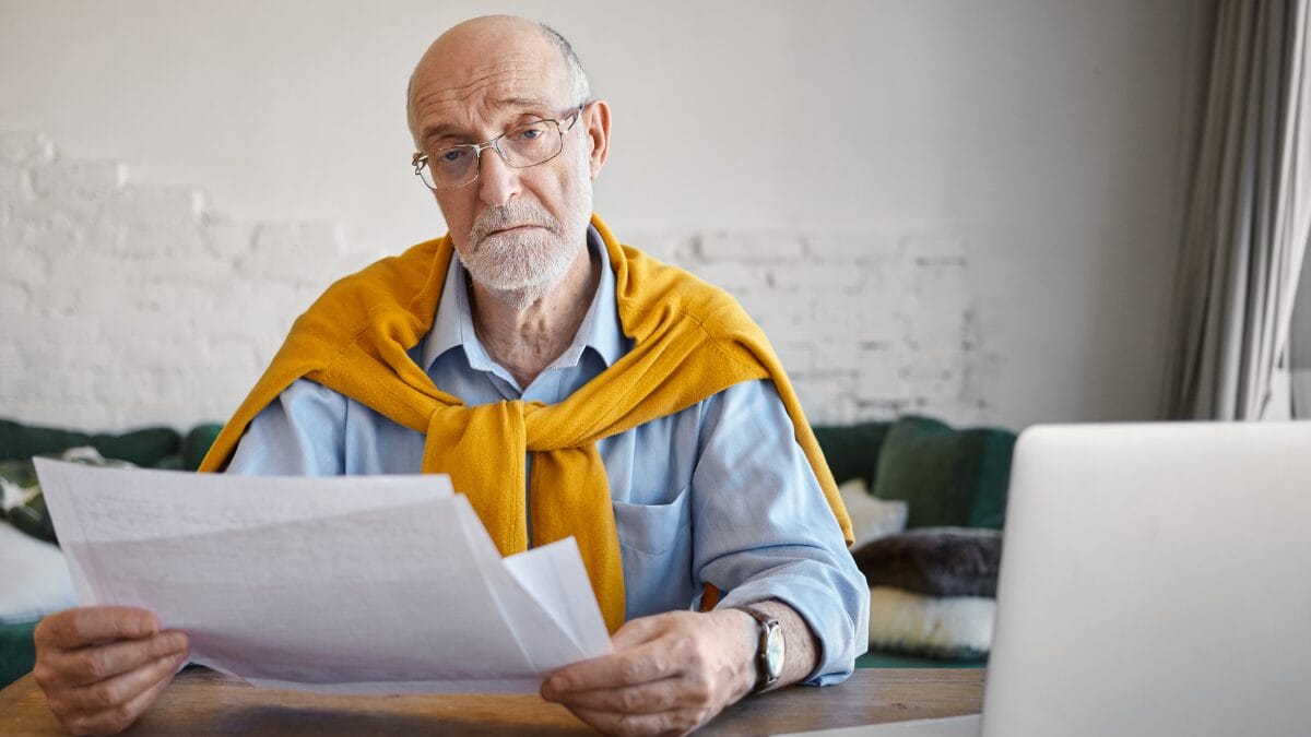 Homem idoso de óculos e suéter amarelo sobre os ombros, sentado à mesa, segura documentos com expressão de preocupação e dúvida. Ao fundo, um ambiente doméstico e um notebook. Imagem ilustra a aplicação da lei contra o superendividamento de idosos.